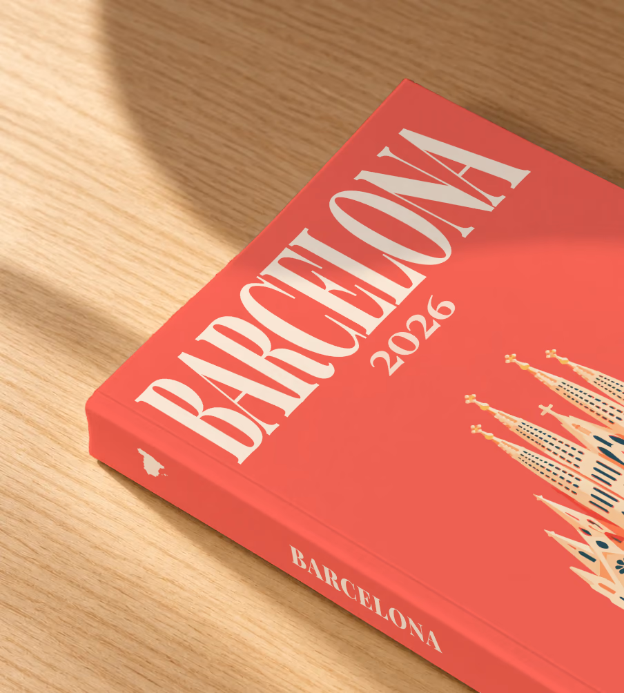 Spain softcover photo book with the Barcelona illustrated cover of the Sagrada Família on coral red, lying on an oak desk in dappled afternoon sunlight
