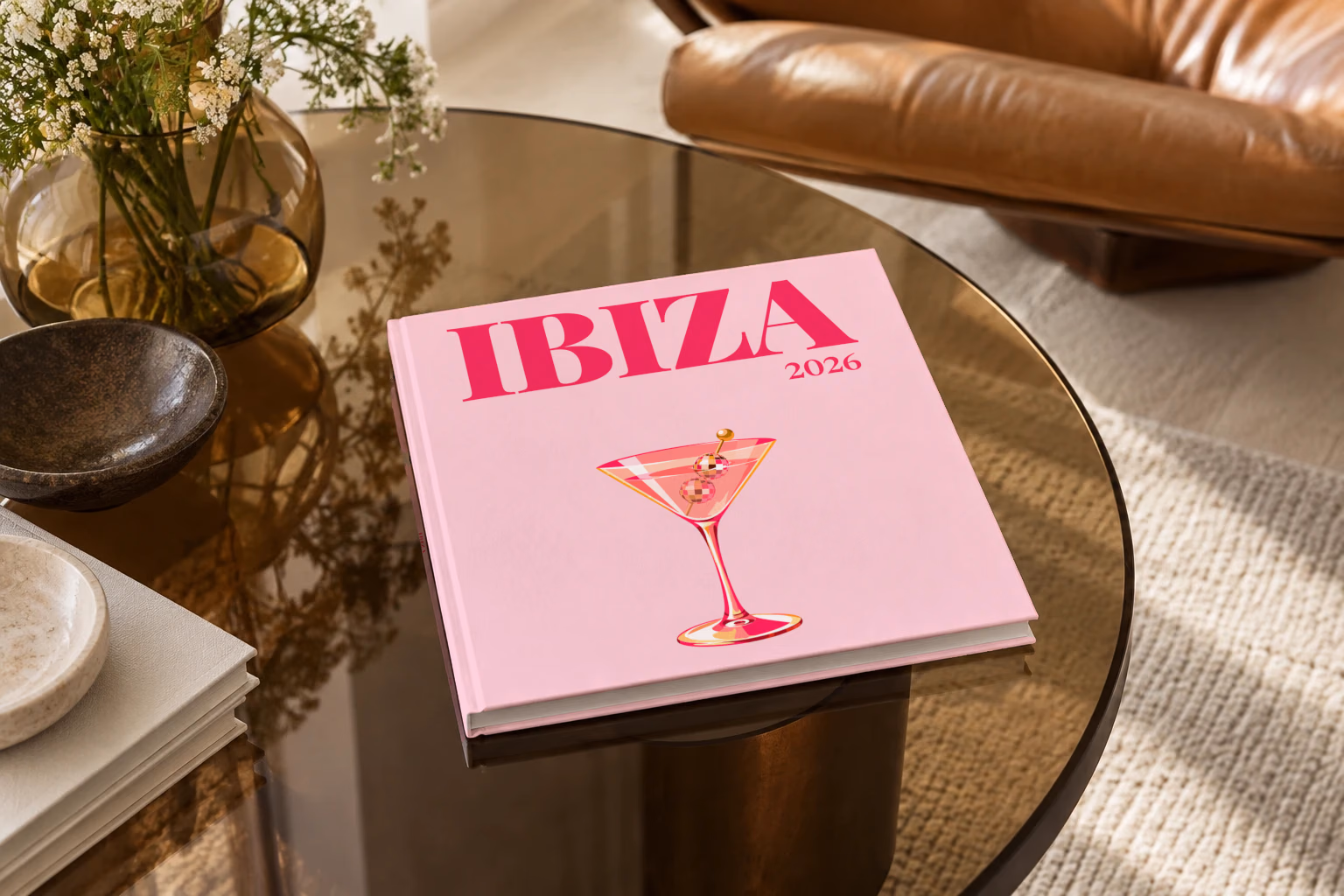 Spain square hardcover photo book with the Ibiza illustrated cover of a martini with a disco-ball garnish on pale pink, on a smoked glass coffee table beside a leather armchair in warm afternoon light