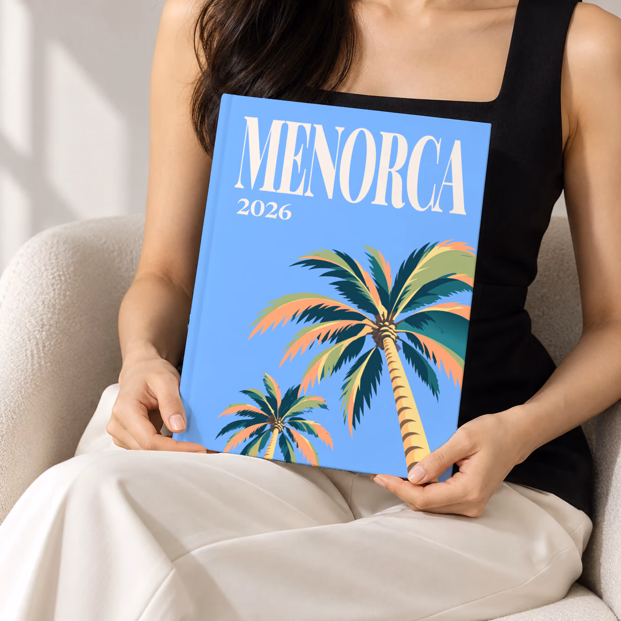Spain hardcover photo book with the Menorca illustrated cover of palm trees on blue, held upright in two hands by a woman in a black top, sitting in a cream boucle armchair