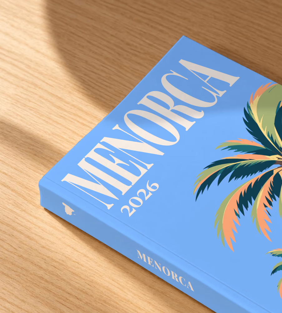 Spain softcover photo book with the Menorca illustrated cover of palm trees on blue, lying on an oak desk in dappled afternoon sunlight