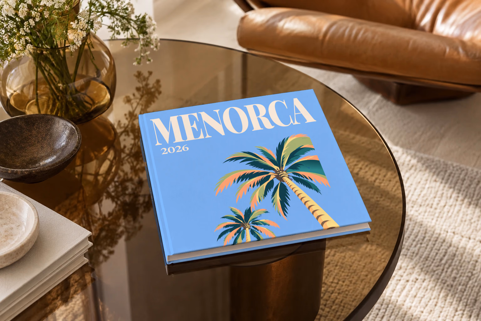 Spain square hardcover photo book with the Menorca illustrated cover of palm trees on blue, on a smoked glass coffee table beside a leather armchair in warm afternoon light