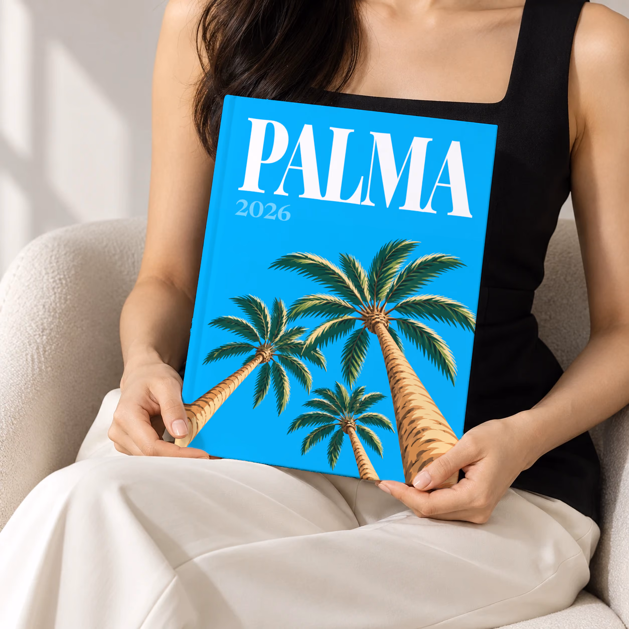 Spain hardcover photo book with the Palma illustrated cover of palm trees on bright blue, held upright in two hands by a woman in a black top, sitting in a cream boucle armchair