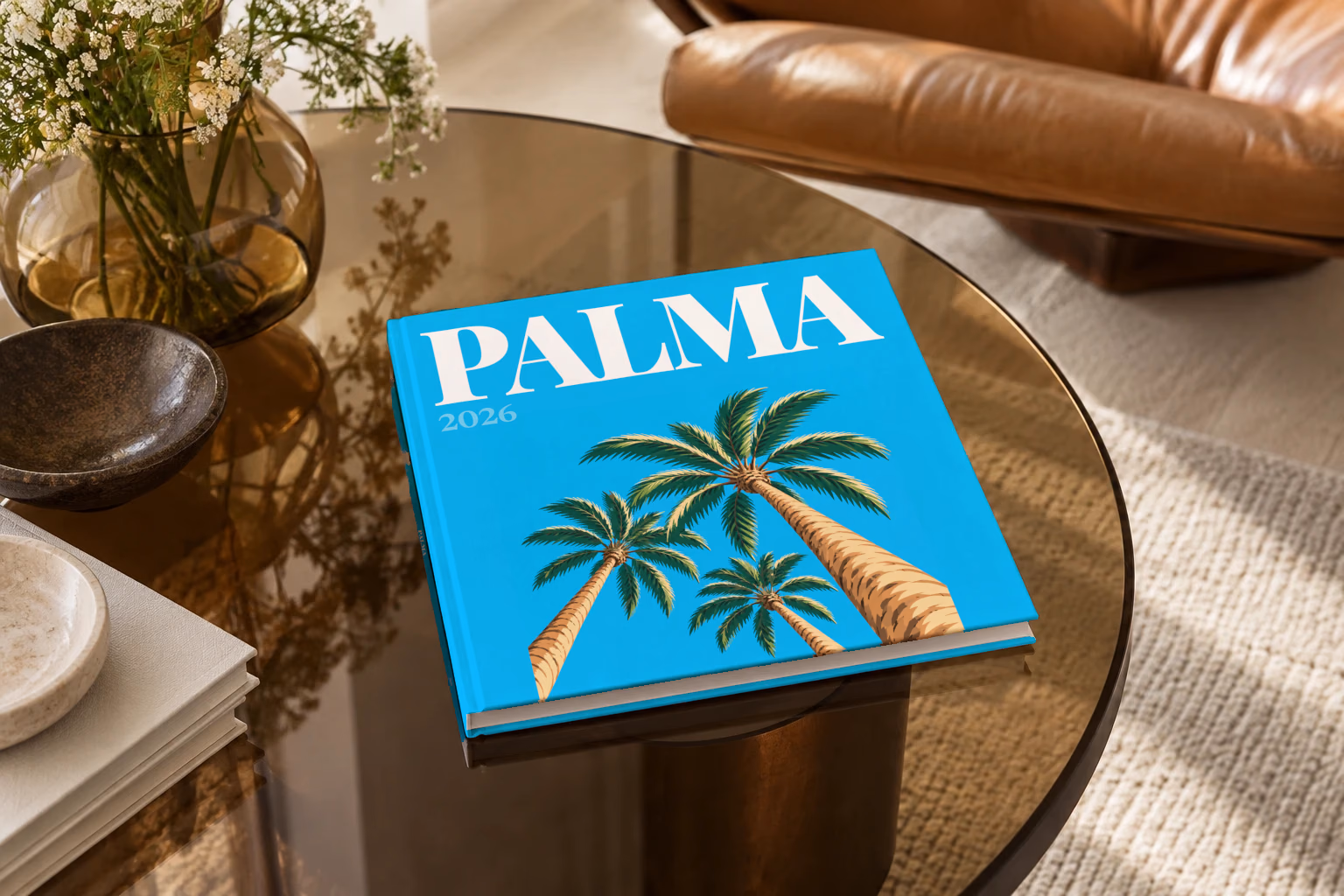 Spain square hardcover photo book with the Palma illustrated cover of palm trees on bright blue, on a smoked glass coffee table beside a leather armchair in warm afternoon light