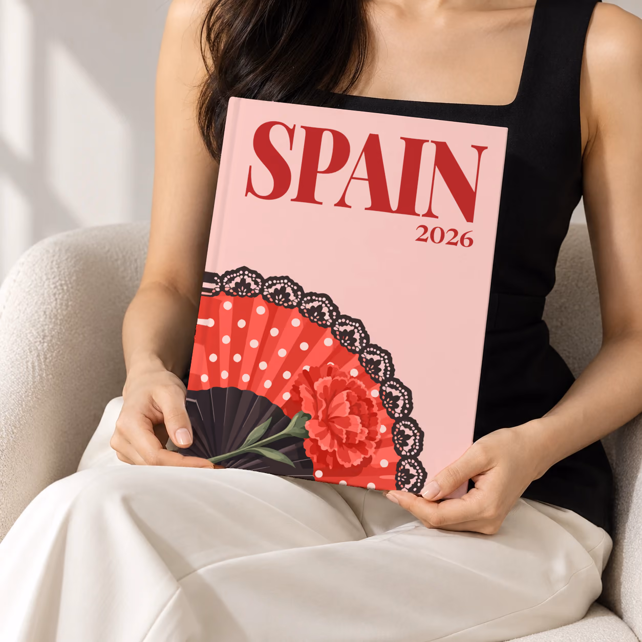 Spain hardcover photo book with the illustrated cover of a polka-dot flamenco fan and a carnation on blush pink, held upright in two hands by a woman in a black top, sitting in a cream boucle armchair