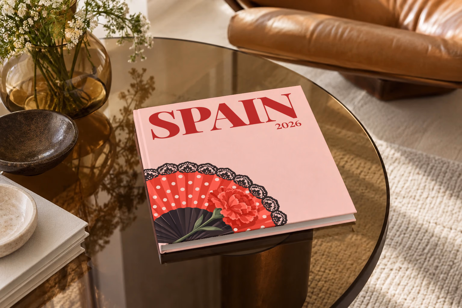 Spain square hardcover photo book with the illustrated cover of a polka-dot flamenco fan and a carnation on blush pink, on a smoked glass coffee table beside a leather armchair in warm afternoon light