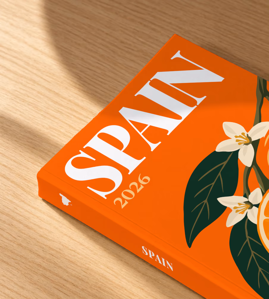 Spain softcover photo book with the illustrated cover of a halved orange and orange blossom on orange, lying on an oak desk in dappled afternoon sunlight