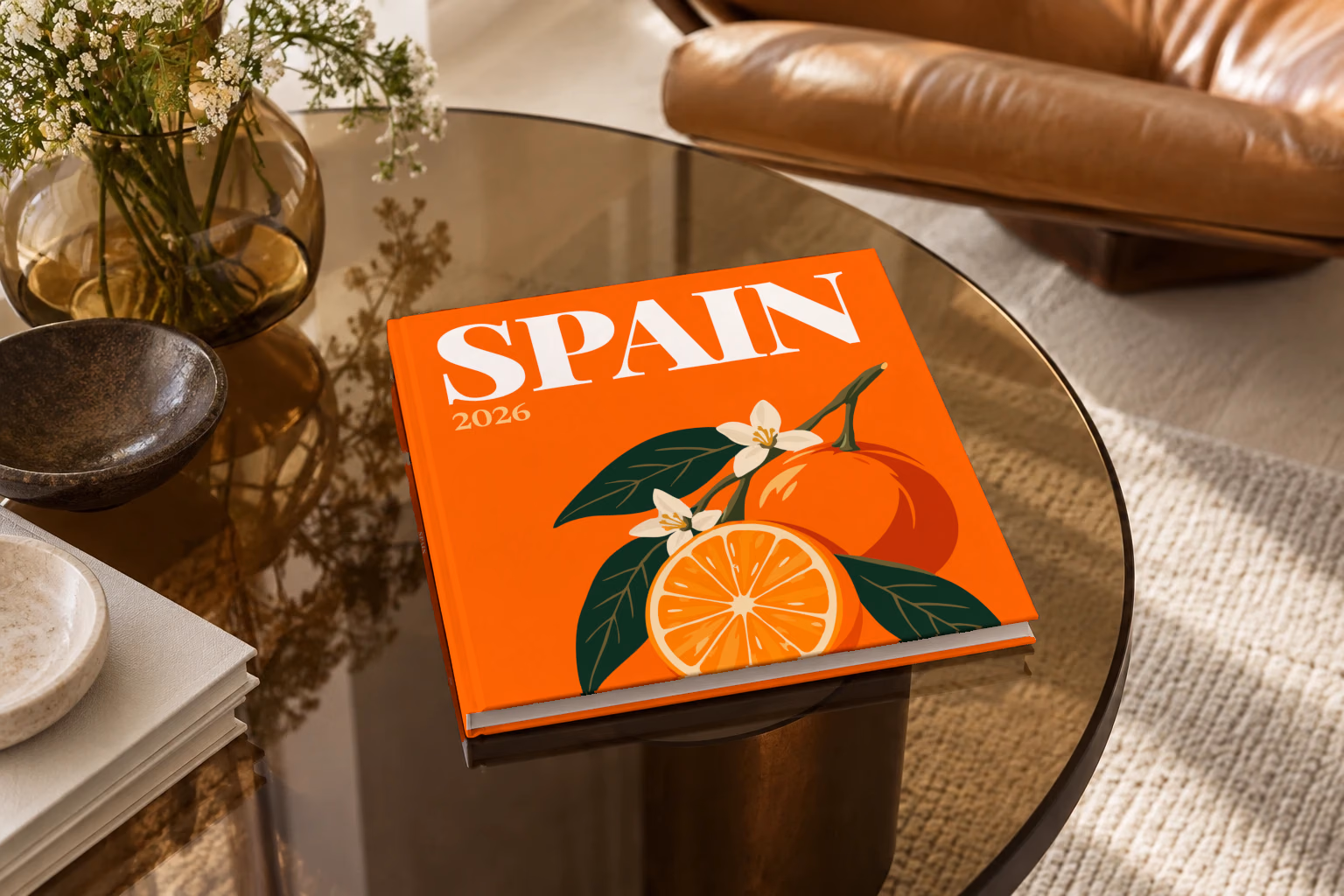 Spain square hardcover photo book with the illustrated cover of a halved orange and orange blossom on orange, on a smoked glass coffee table beside a leather armchair in warm afternoon light