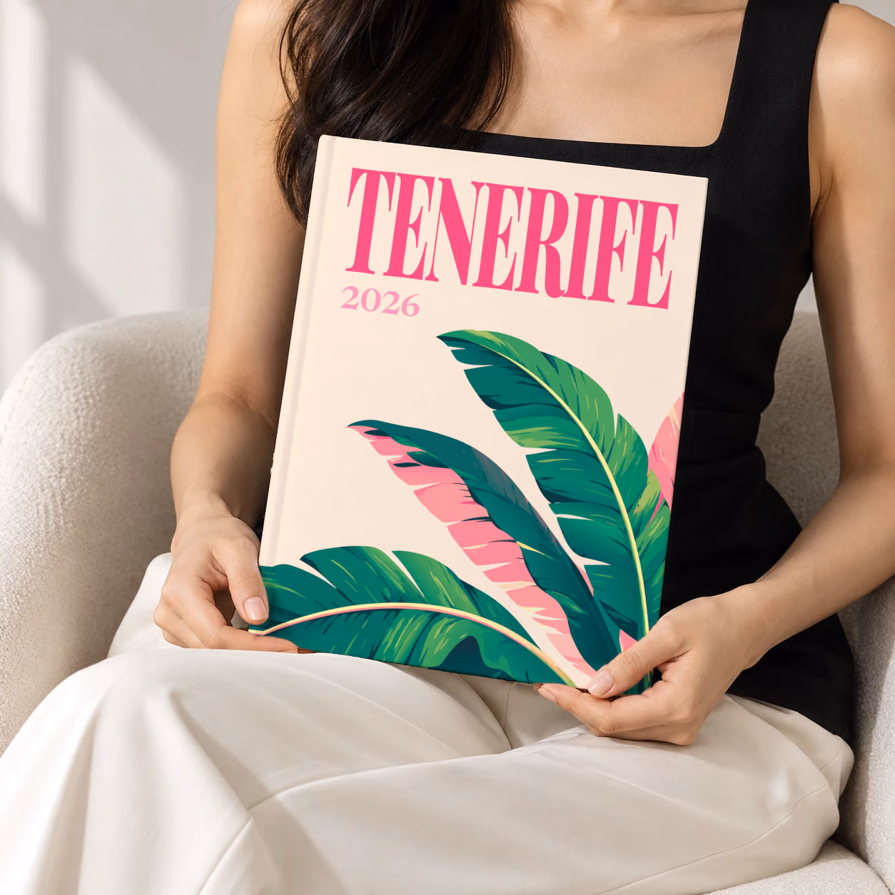 Spain hardcover photo book with the Tenerife illustrated cover of banana leaves on cream, held upright in two hands by a woman in a black top, sitting in a cream boucle armchair