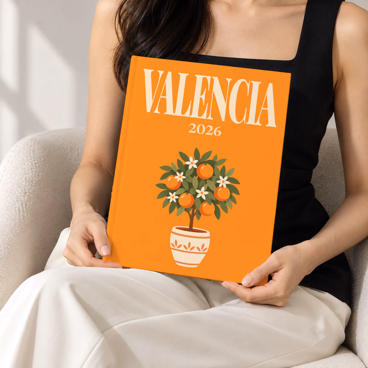 Spain hardcover photo book with the Valencia illustrated cover of a potted orange tree on orange, held upright in two hands by a woman in a black top, sitting in a cream boucle armchair