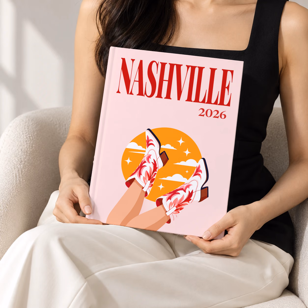 Tennessee hardcover photo book with the Nashville illustrated cover of kicking legs in red and white cowboy boots against a yellow sun on pale pink, held upright in two hands by a woman in a black top, sitting in a cream boucle armchair