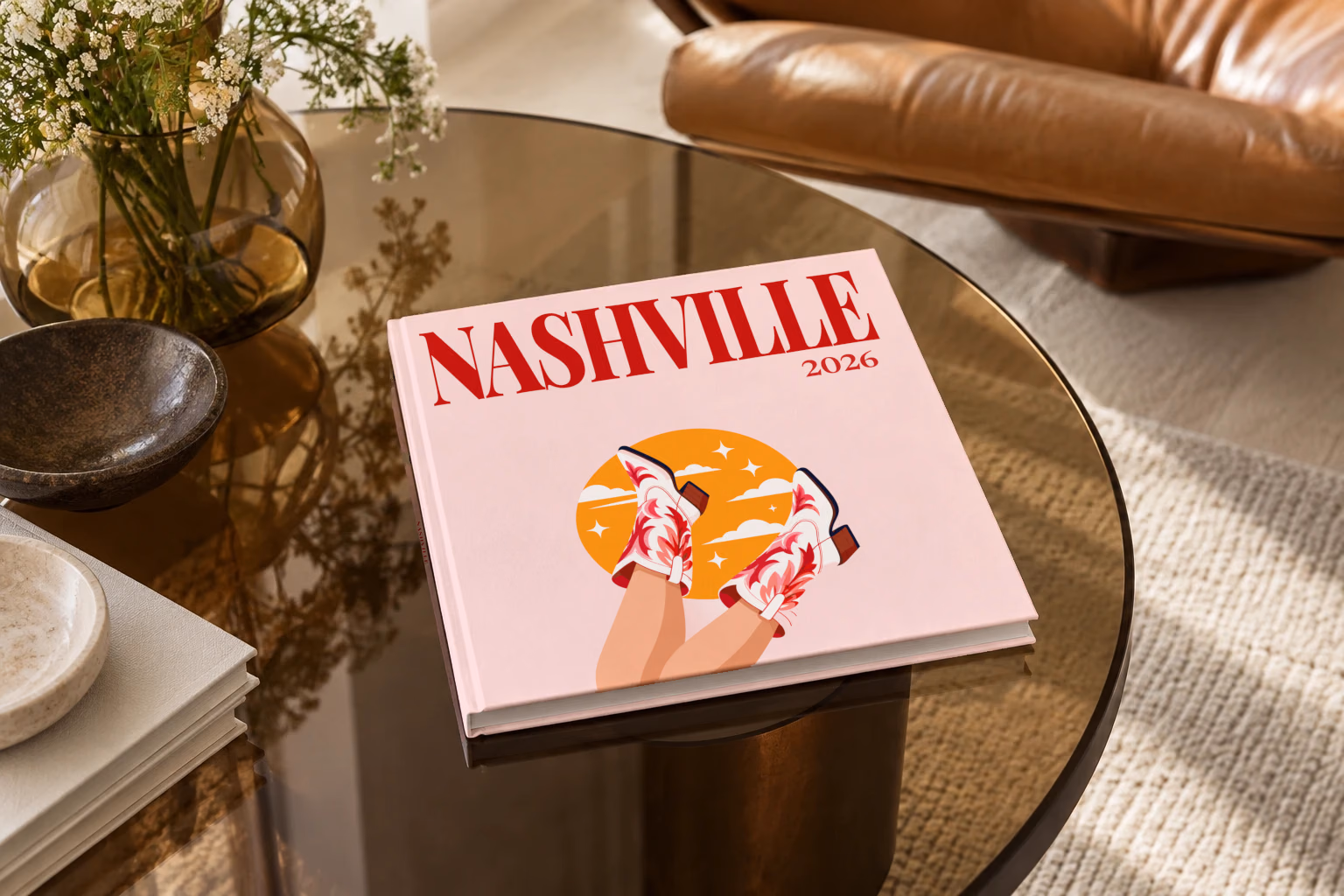 Tennessee square hardcover photo book with the Nashville illustrated cover of kicking legs in red and white cowboy boots against a yellow sun on pale pink, on a smoked glass coffee table beside a leather armchair in warm afternoon light