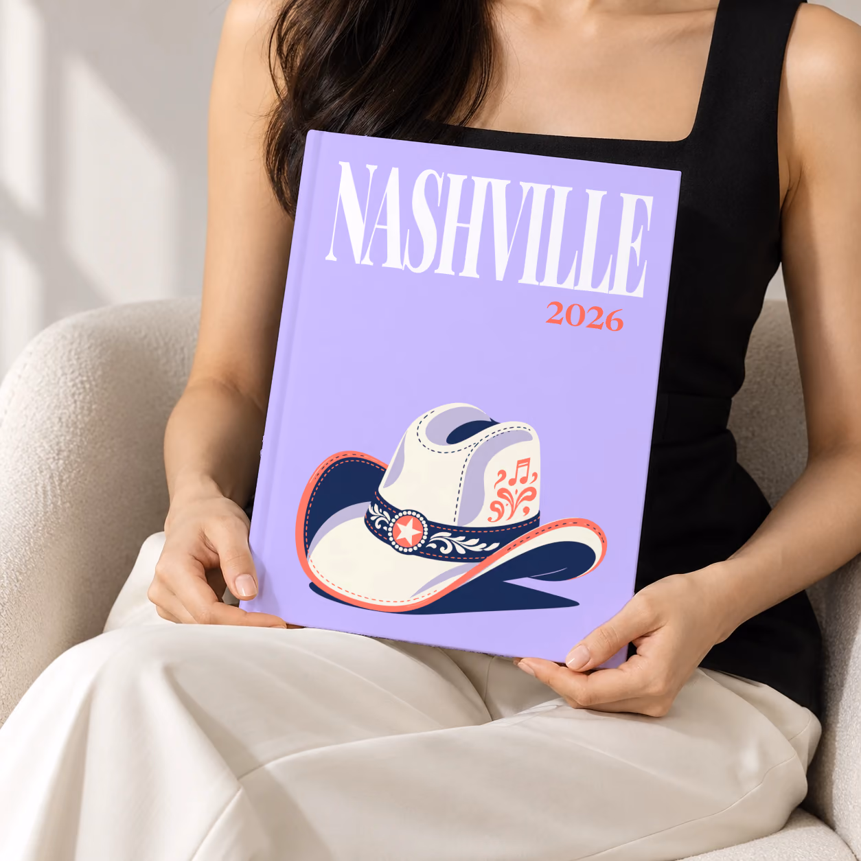 Tennessee hardcover photo book with the Nashville illustrated cover of a white cowboy hat with music notes on lavender, held upright in two hands by a woman in a black top, sitting in a cream boucle armchair