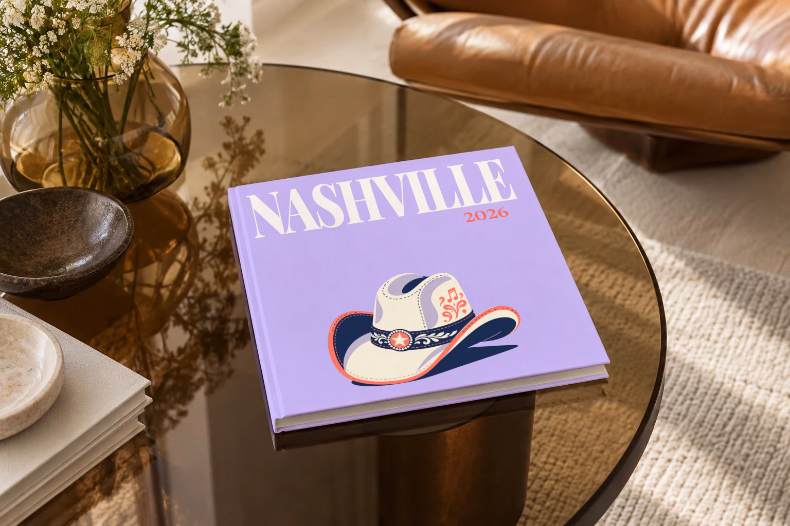 Tennessee square hardcover photo book with the Nashville illustrated cover of a white cowboy hat with music notes on lavender, on a smoked glass coffee table beside a leather armchair in warm afternoon light