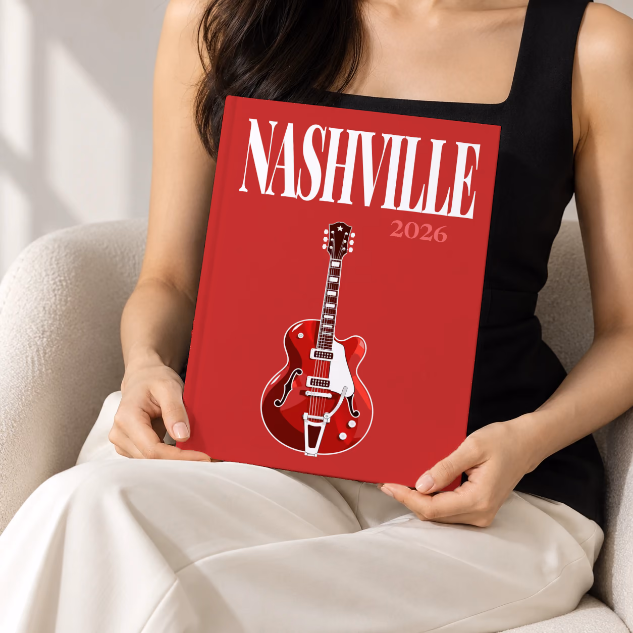 Tennessee hardcover photo book with the Nashville illustrated cover of a red electric guitar on crimson, held upright in two hands by a woman in a black top, sitting in a cream boucle armchair