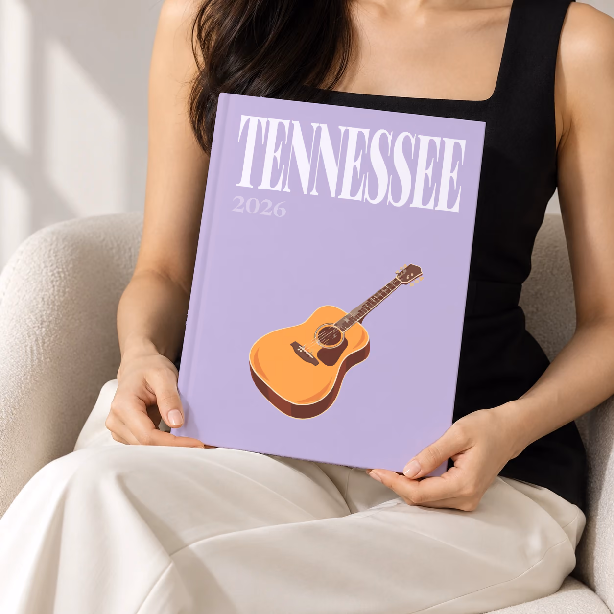 Tennessee hardcover photo book with the illustrated cover of an acoustic guitar on lilac, held upright in two hands by a woman in a black top, sitting in a cream boucle armchair