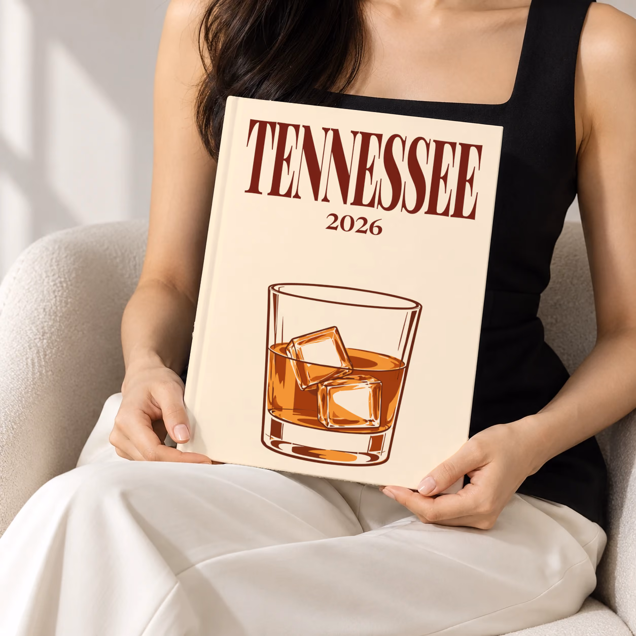 Tennessee hardcover photo book with the illustrated cover of a glass of Tennessee whiskey on the rocks on pale apricot, held upright in two hands by a woman in a black top, sitting in a cream boucle armchair