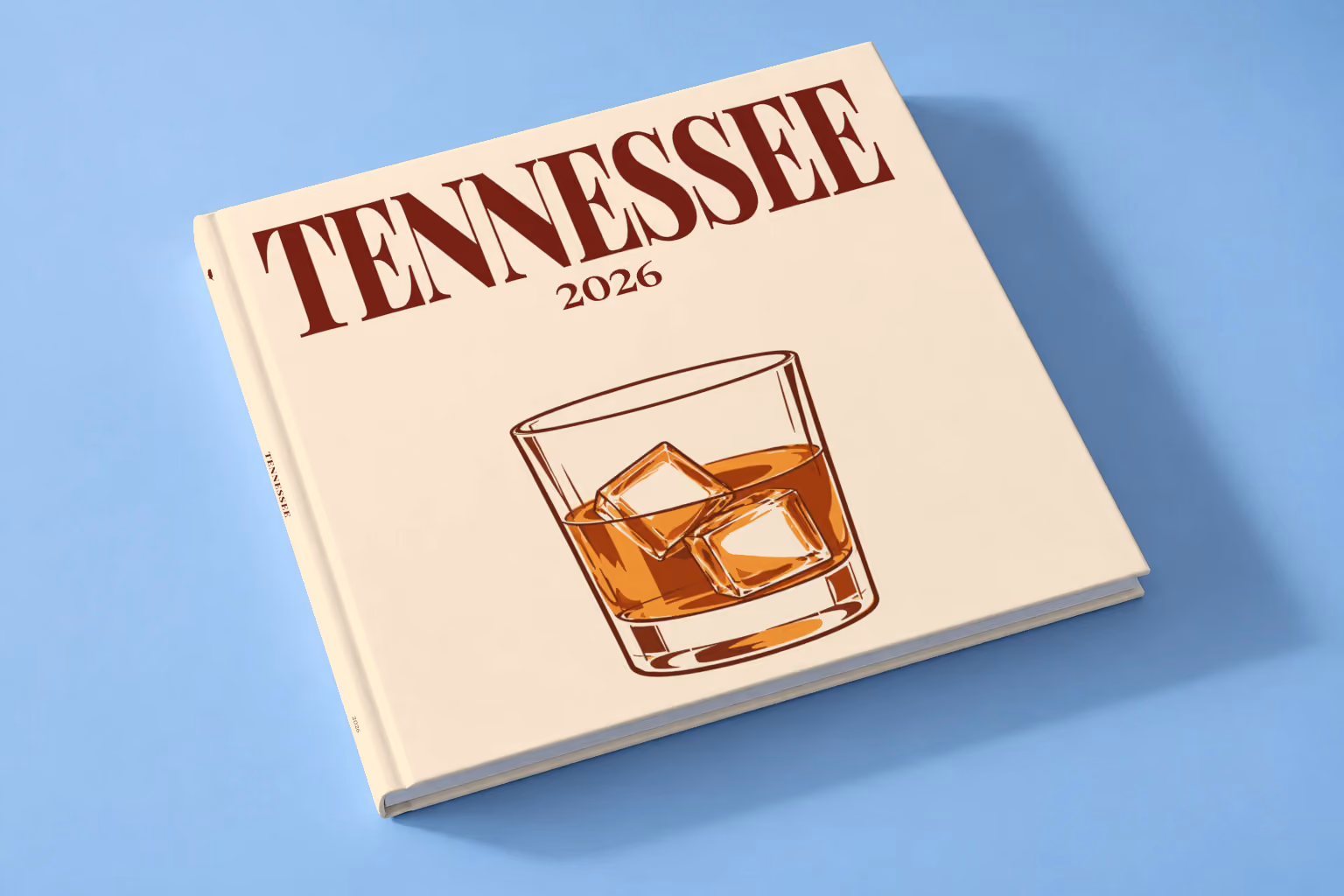 Tennessee square hardcover photo book with the illustrated cover of a glass of Tennessee whiskey on the rocks on pale apricot, lying on a pale blue studio backdrop