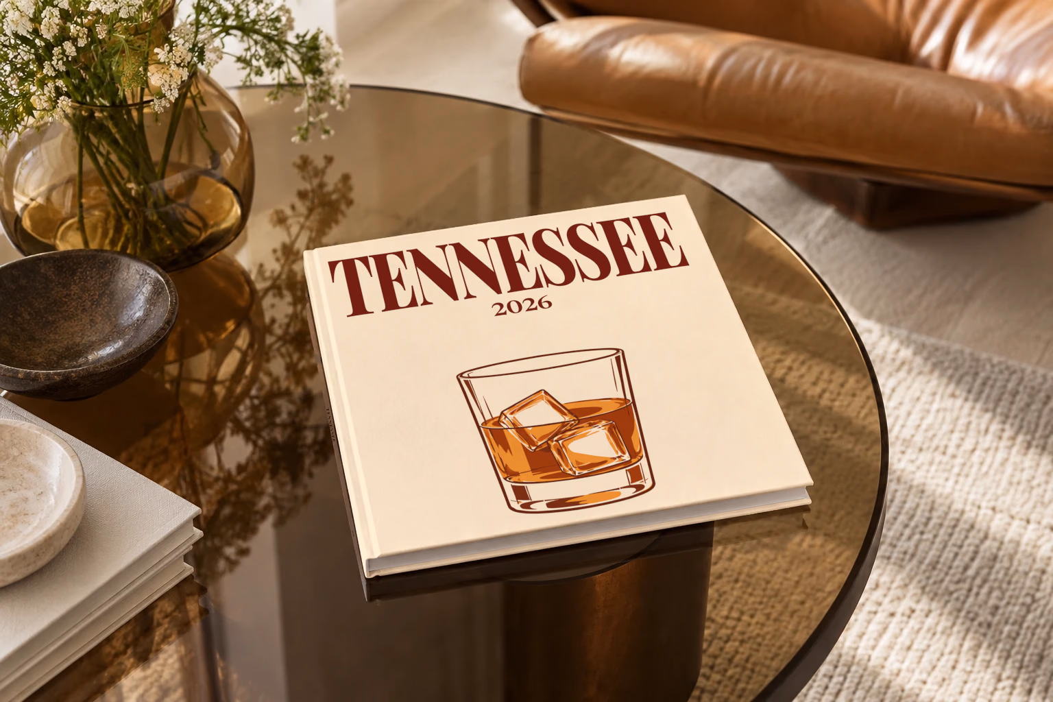 Tennessee square hardcover photo book with the illustrated cover of a glass of Tennessee whiskey on the rocks on pale apricot, on a smoked glass coffee table beside a leather armchair in warm afternoon light