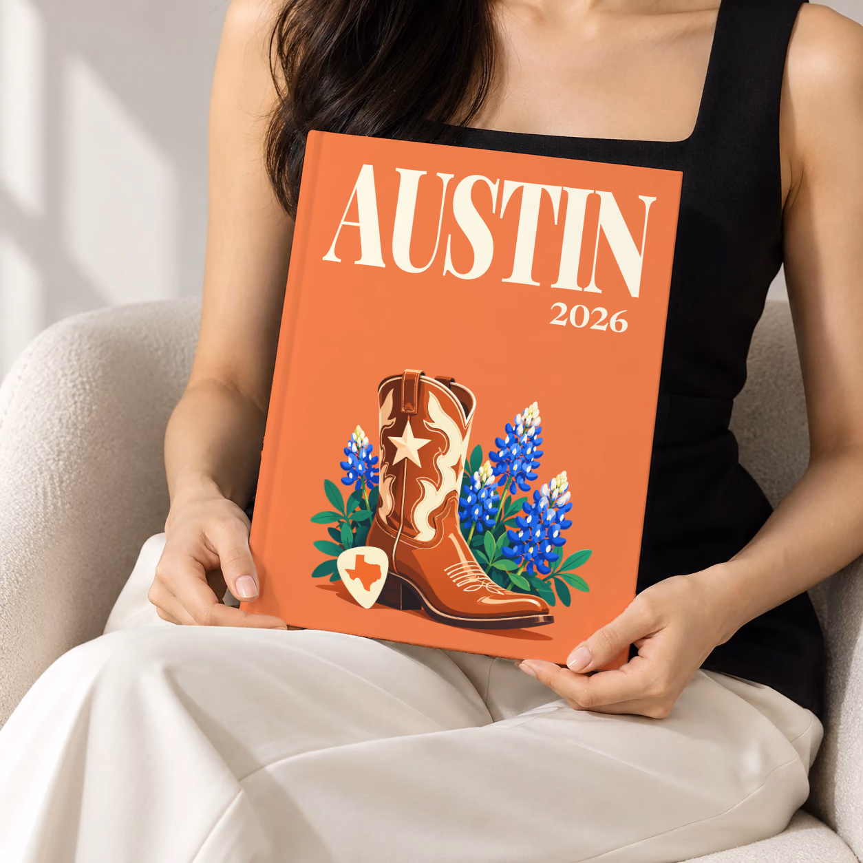 Texas hardcover photo book with the Austin illustrated cover of a cowboy boot with Texas bluebonnets and a guitar pick on coral, held upright in two hands by a woman in a black top, sitting in a cream boucle armchair