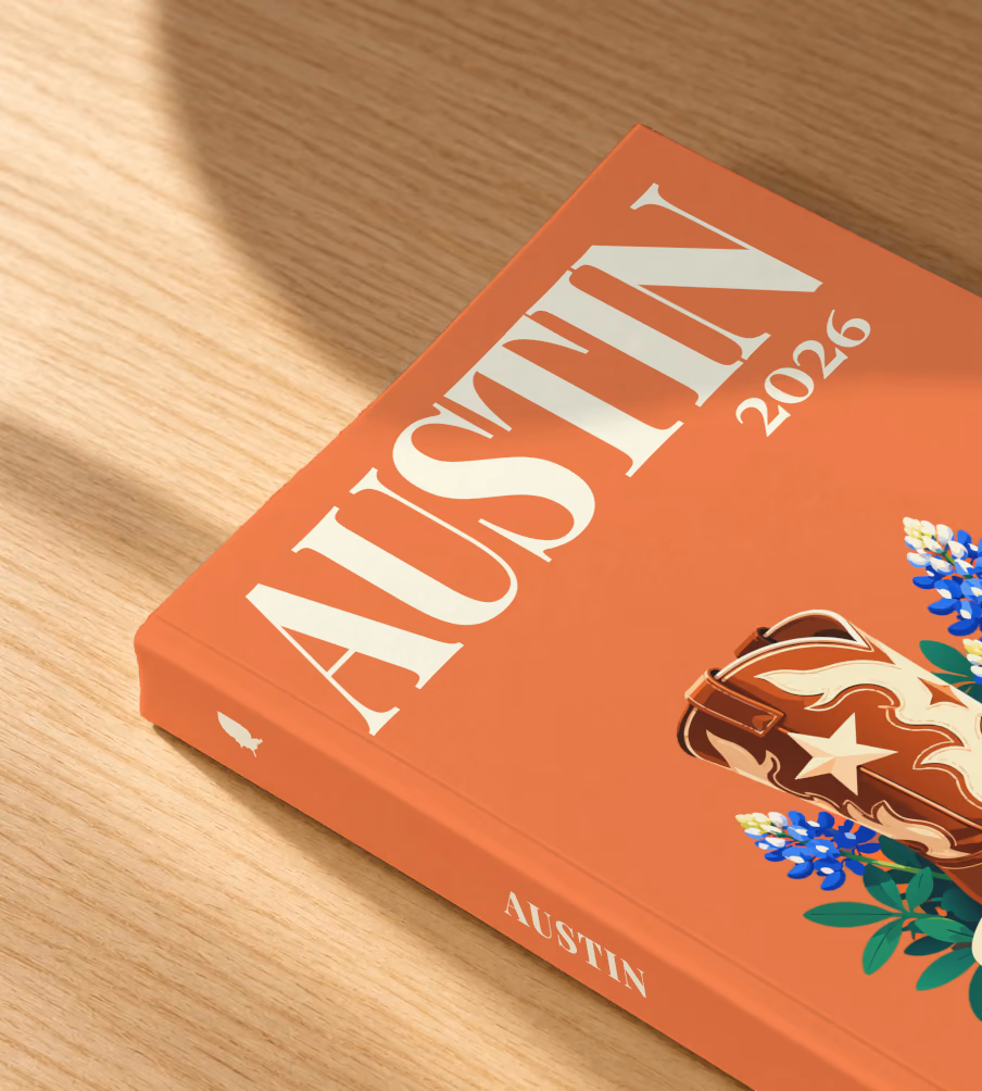 Texas softcover photo book with the Austin illustrated cover of a cowboy boot with Texas bluebonnets and a guitar pick on coral, lying on an oak desk in dappled afternoon sunlight