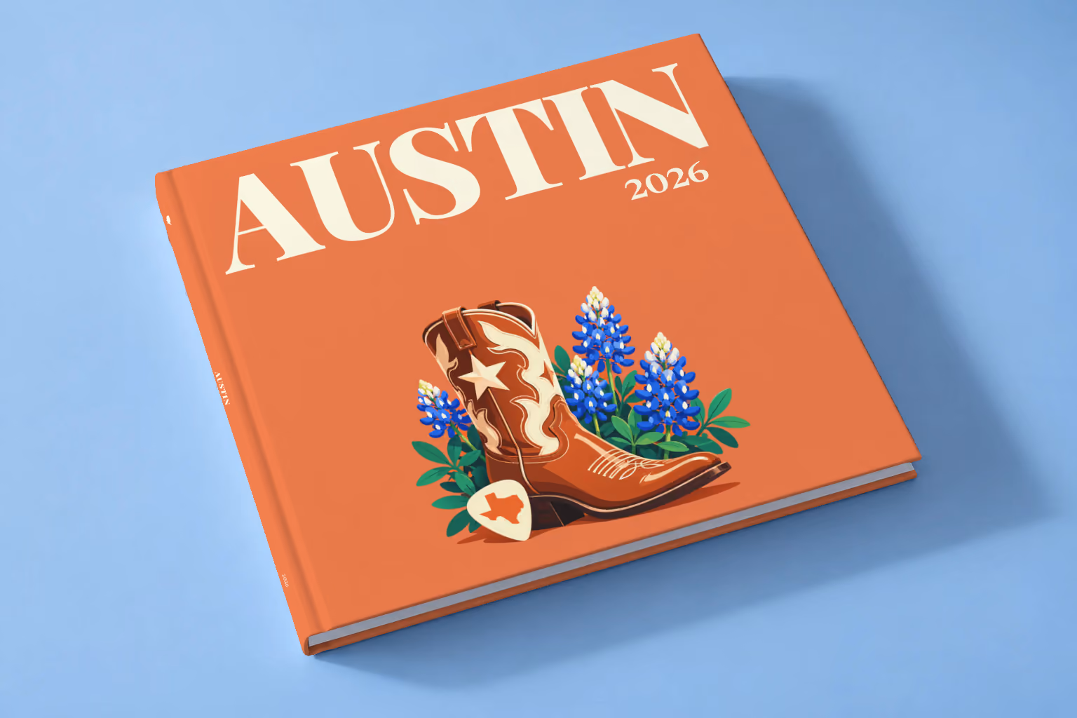 Texas square hardcover photo book with the Austin illustrated cover of a cowboy boot with Texas bluebonnets and a guitar pick on coral, lying on a pale blue studio backdrop