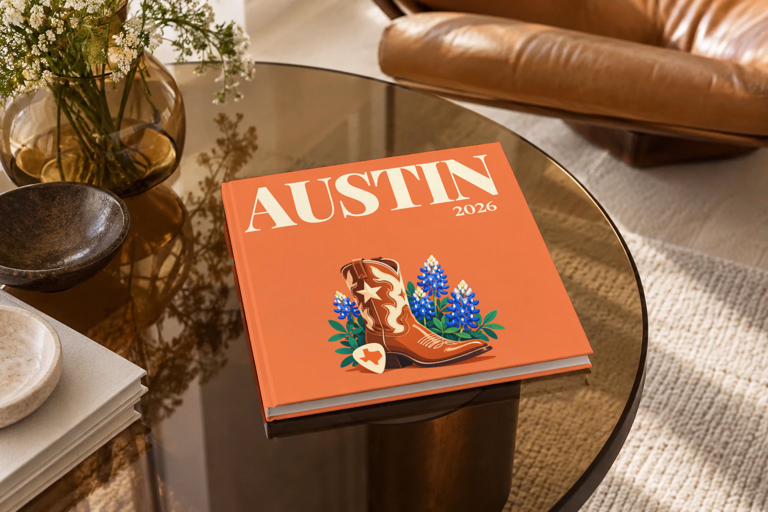 Texas square hardcover photo book with the Austin illustrated cover of a cowboy boot with Texas bluebonnets and a guitar pick on coral, on a smoked glass coffee table beside a leather armchair in warm afternoon light