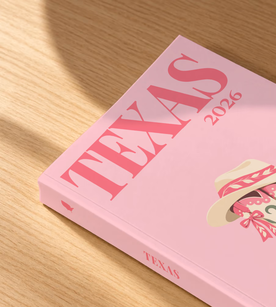 Texas softcover photo book with the illustrated cover of pink cowboy boots with bows and a cowboy hat on blush pink, lying on an oak desk in dappled afternoon sunlight