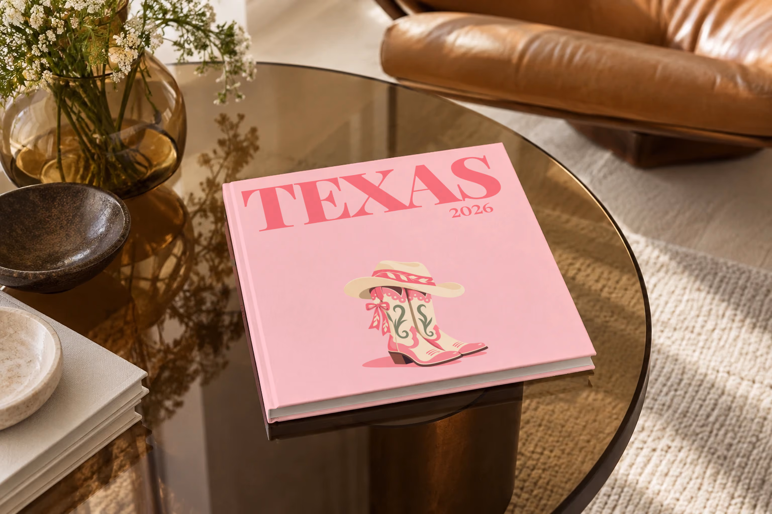 Texas square hardcover photo book with the illustrated cover of pink cowboy boots with bows and a cowboy hat on blush pink, on a smoked glass coffee table beside a leather armchair in warm afternoon light