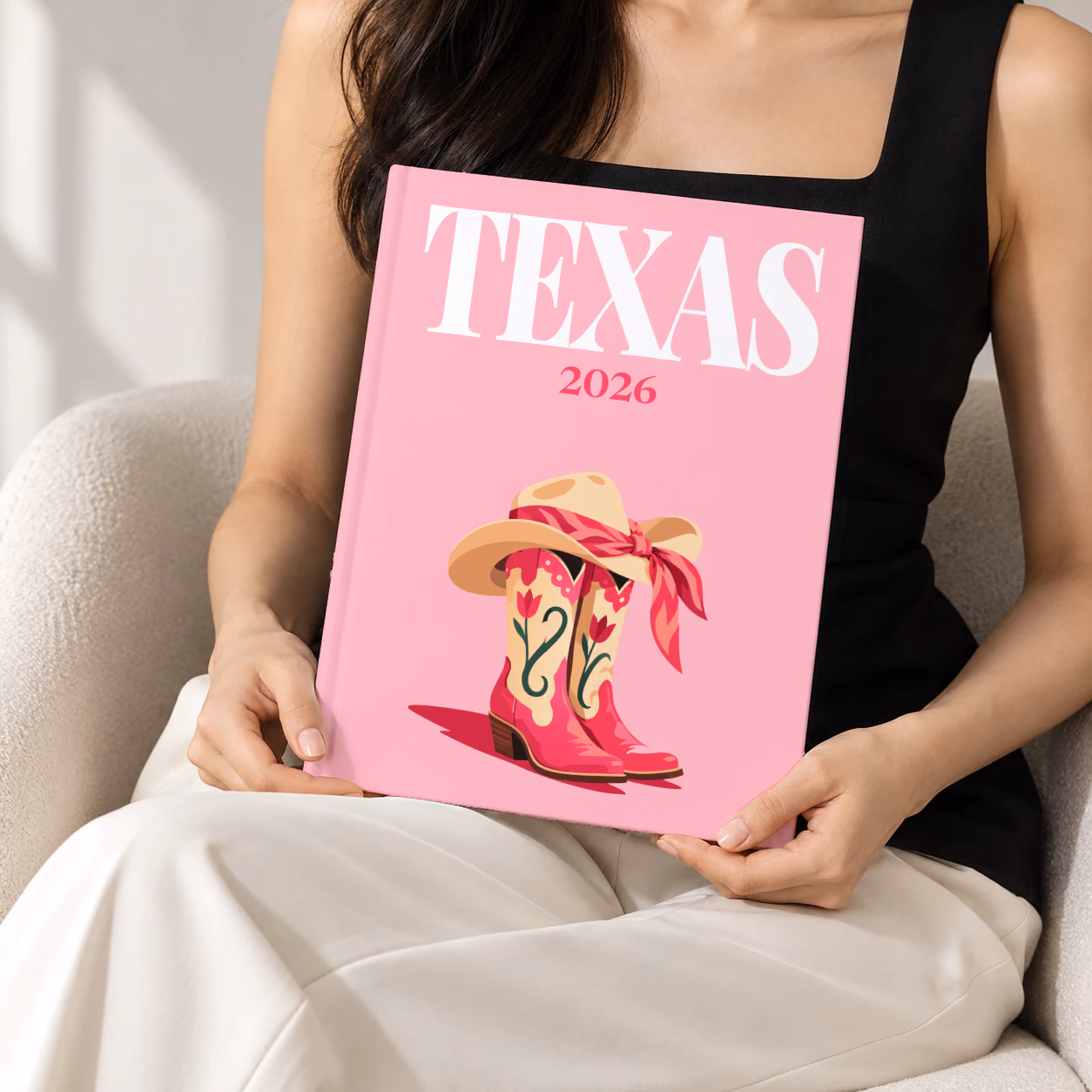 Texas hardcover photo book with the illustrated cover of pink cowboy boots and a cowboy hat with a pink bandana on pink, held upright in two hands by a woman in a black top, sitting in a cream boucle armchair