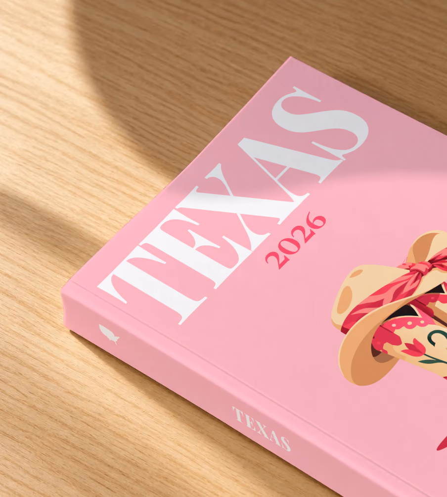 Texas softcover photo book with the illustrated cover of pink cowboy boots and a cowboy hat with a pink bandana on pink, lying on an oak desk in dappled afternoon sunlight