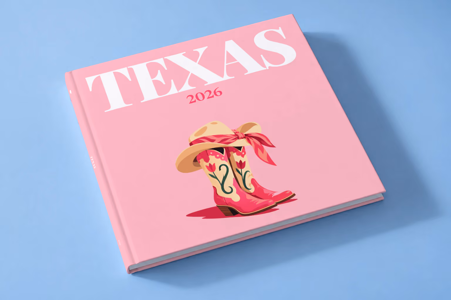 Texas square hardcover photo book with the illustrated cover of pink cowboy boots and a cowboy hat with a pink bandana on pink, lying on a pale blue studio backdrop