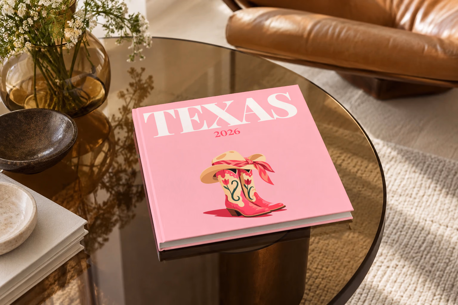 Texas square hardcover photo book with the illustrated cover of pink cowboy boots and a cowboy hat with a pink bandana on pink, on a smoked glass coffee table beside a leather armchair in warm afternoon light