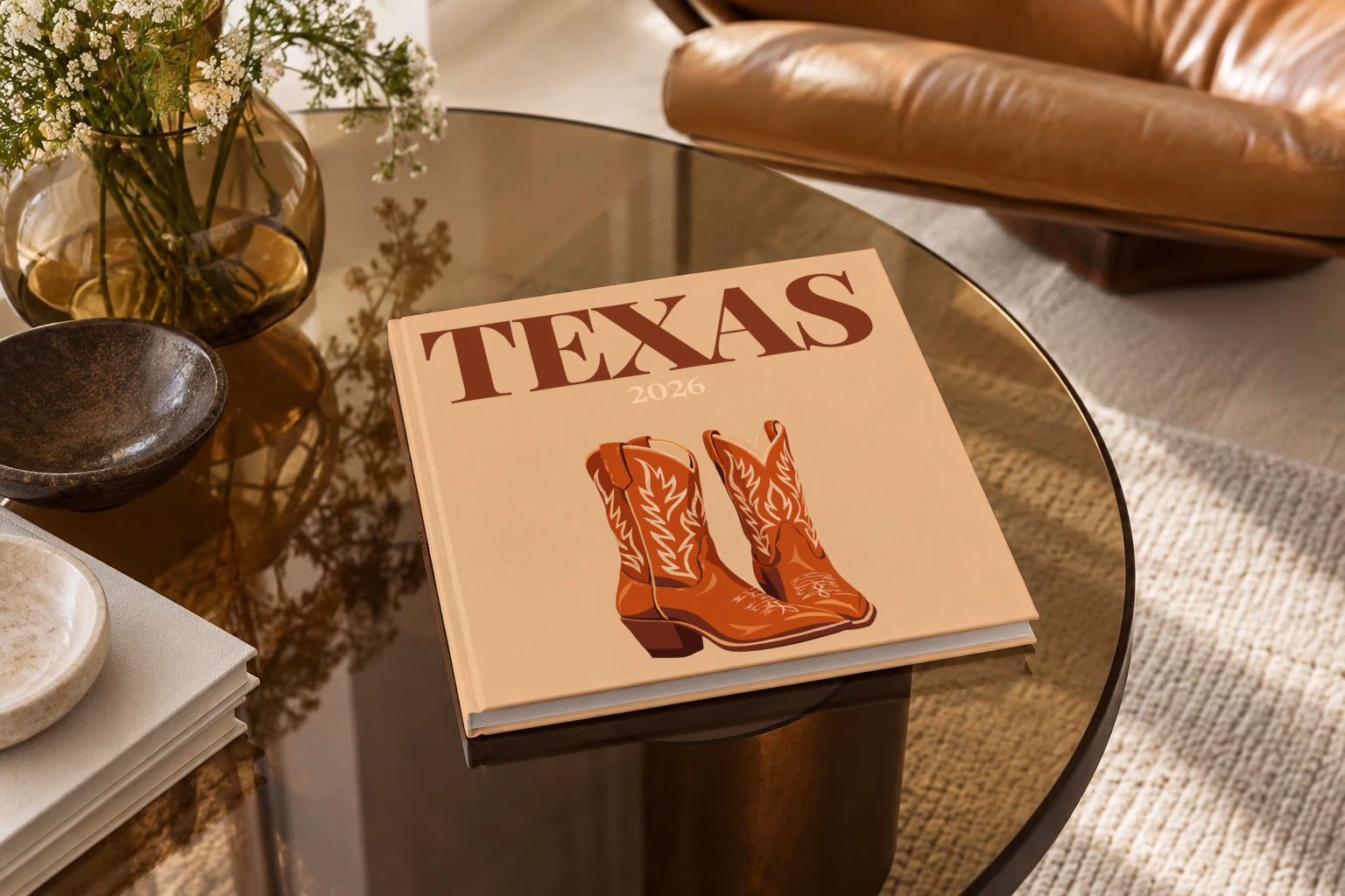 Texas square hardcover photo book with the illustrated cover of a pair of brown leather cowboy boots on tan, on a smoked glass coffee table beside a leather armchair in warm afternoon light