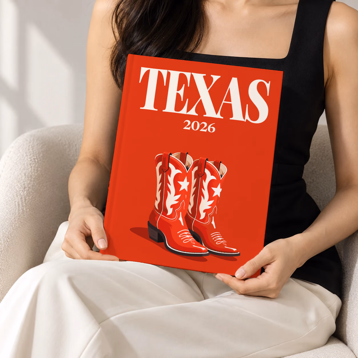 Texas hardcover photo book with the illustrated cover of a pair of red cowboy boots with white stars on red, held upright in two hands by a woman in a black top, sitting in a cream boucle armchair