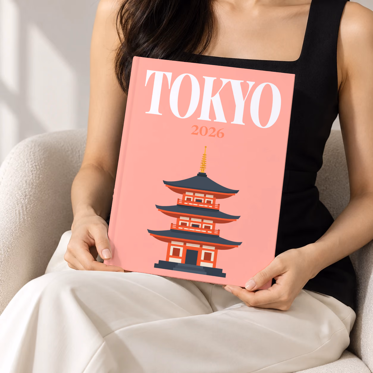 Tokyo hardcover photo book with the illustrated cover of a pagoda on salmon pink, held upright in two hands by a woman in a black top, sitting in a cream boucle armchair