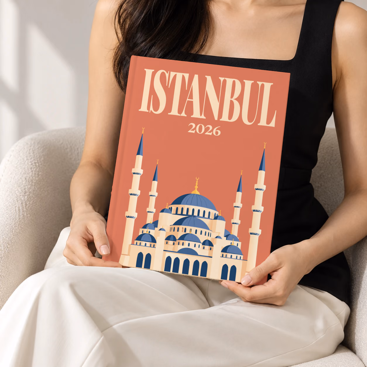 Turkey hardcover photo book with the Istanbul illustrated cover of the domes and minarets of the Blue Mosque on terracotta, held upright in two hands by a woman in a black top, sitting in a cream boucle armchair