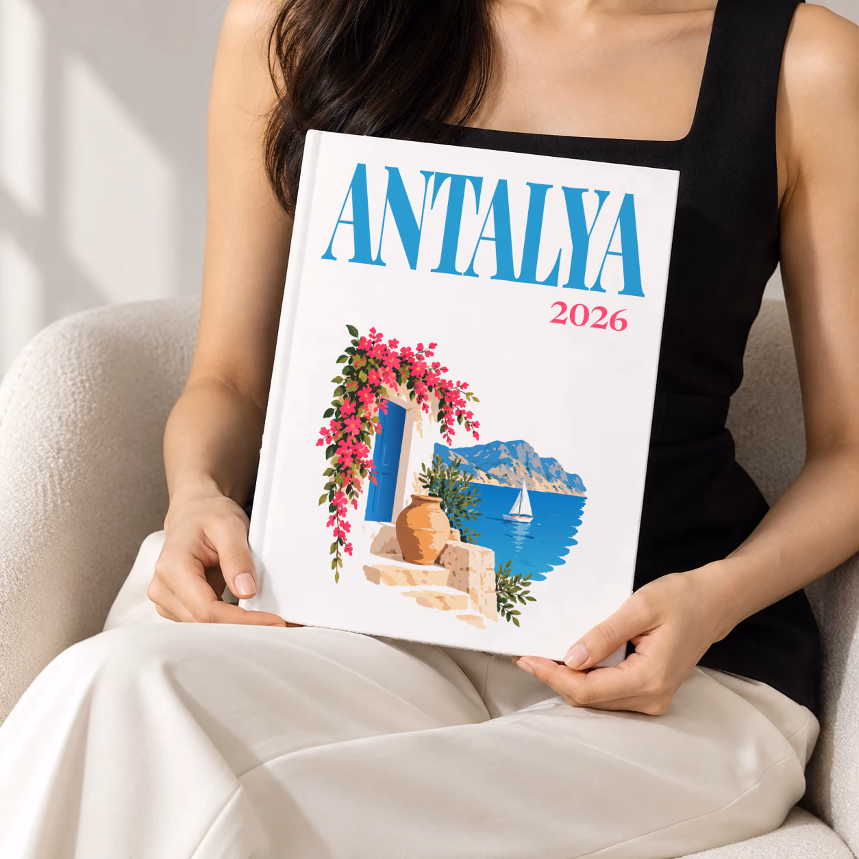 Turkey hardcover photo book with the Antalya illustrated cover of a blue door draped in pink bougainvillea above the Antalya coast on white, held upright in two hands by a woman in a black top, sitting in a cream boucle armchair