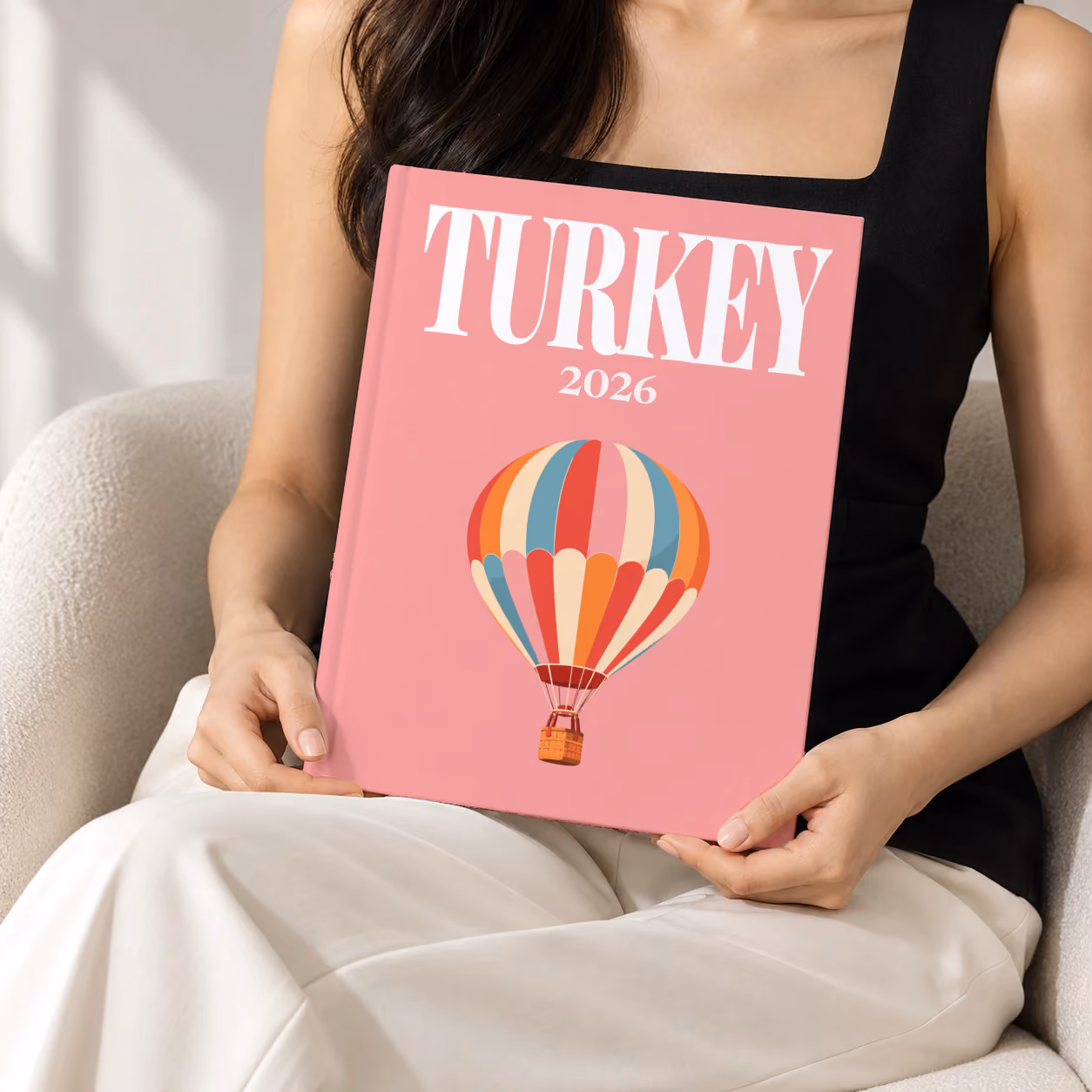 Turkey hardcover photo book with the illustrated cover of a striped Cappadocia hot air balloon on pink, held upright in two hands by a woman in a black top, sitting in a cream boucle armchair