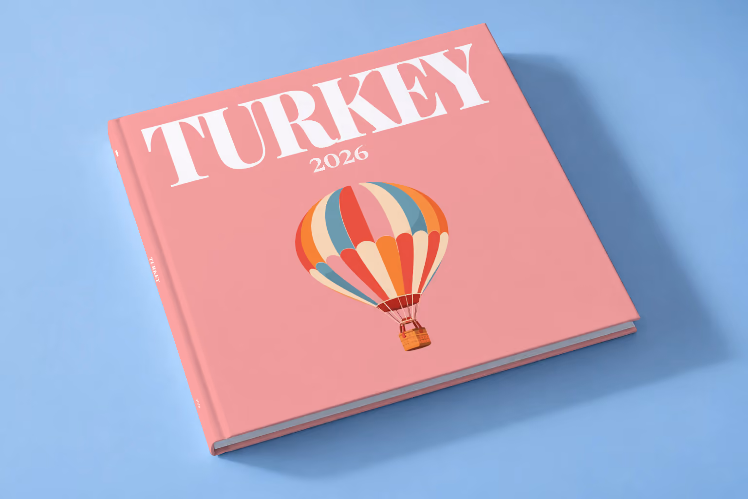 Turkey square hardcover photo book with the illustrated cover of a striped Cappadocia hot air balloon on pink, lying on a pale blue studio backdrop
