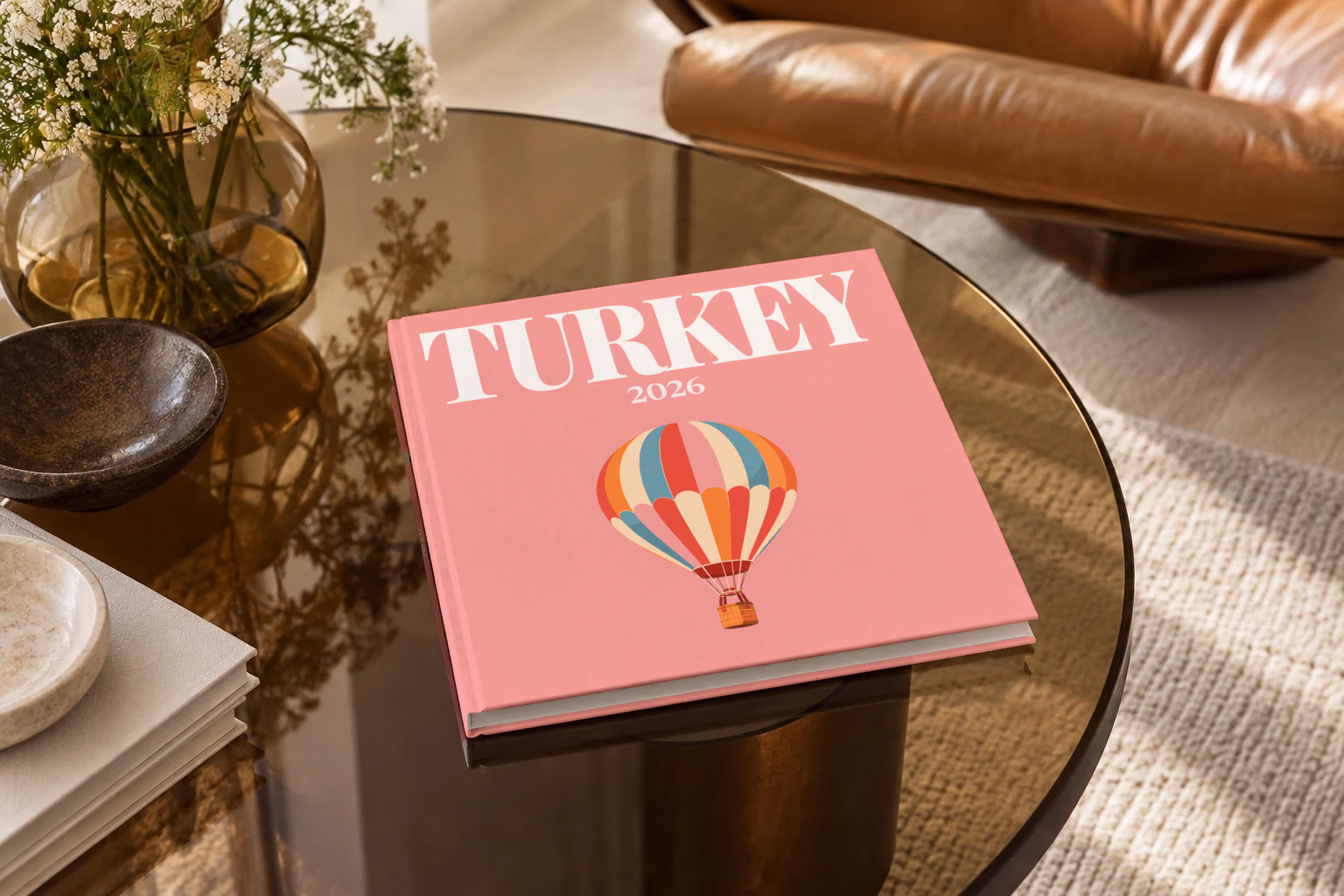 Turkey square hardcover photo book with the illustrated cover of a striped Cappadocia hot air balloon on pink, on a smoked glass coffee table beside a leather armchair in warm afternoon light