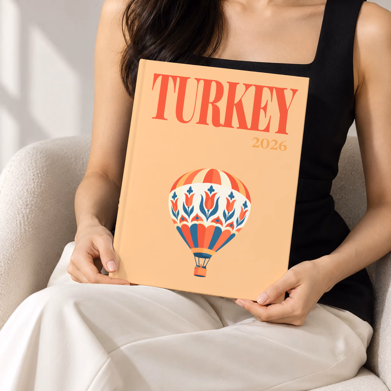 Turkey hardcover photo book with the illustrated cover of a hot air balloon patterned with Turkish tulips on peach, held upright in two hands by a woman in a black top, sitting in a cream boucle armchair