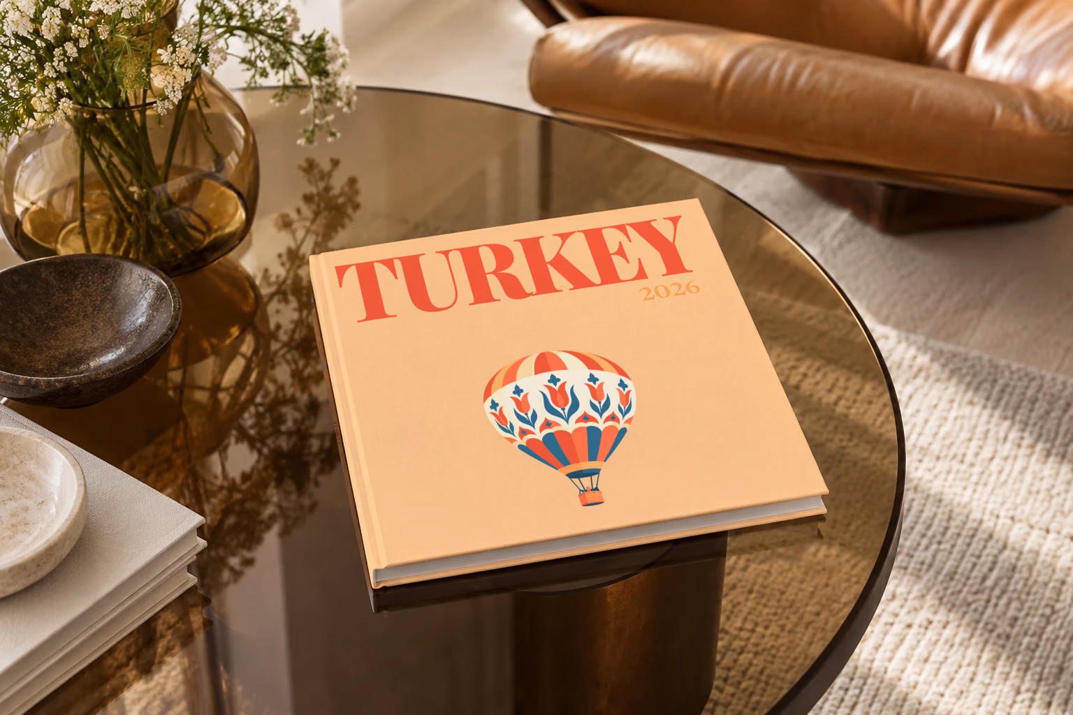 Turkey square hardcover photo book with the illustrated cover of a hot air balloon patterned with Turkish tulips on peach, on a smoked glass coffee table beside a leather armchair in warm afternoon light