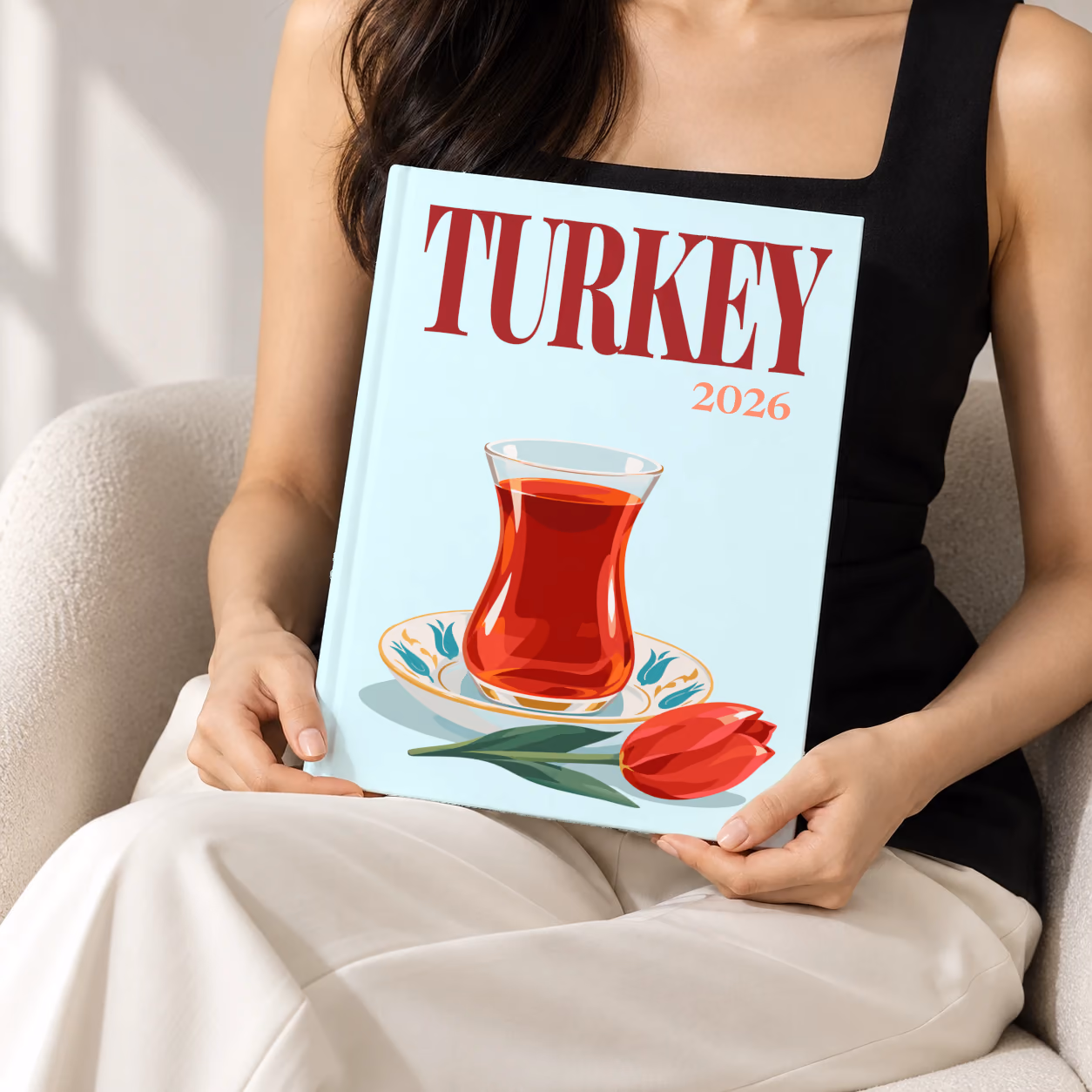 Turkey hardcover photo book with the illustrated cover of a tulip glass of Turkish tea on a saucer with a red tulip on pale aqua, held upright in two hands by a woman in a black top, sitting in a cream boucle armchair