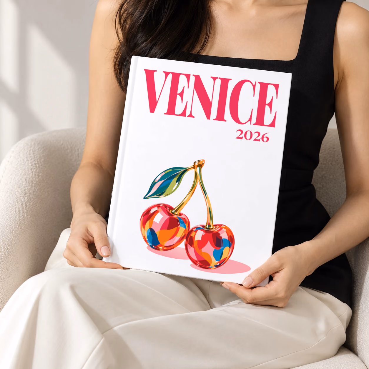 Venice hardcover photo book with the illustrated cover of glass cherries on white, held upright in two hands by a woman in a black top, sitting in a cream boucle armchair