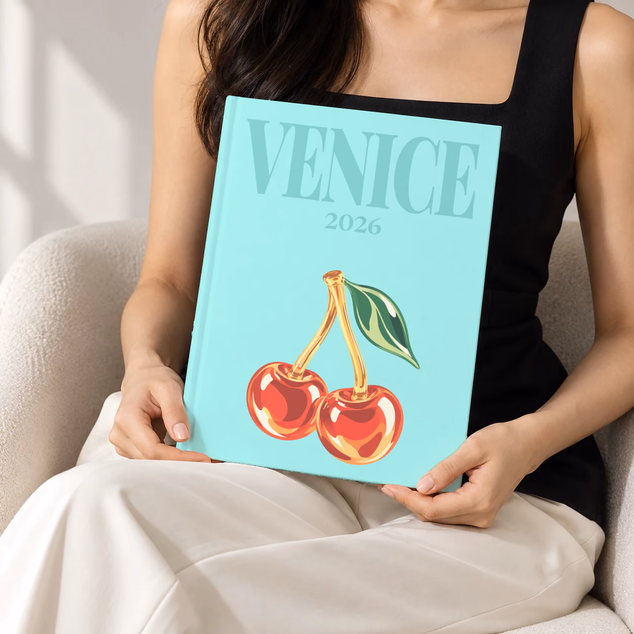 Venice hardcover photo book with the illustrated cover of glass cherries on pale aqua, held upright in two hands by a woman in a black top, sitting in a cream boucle armchair