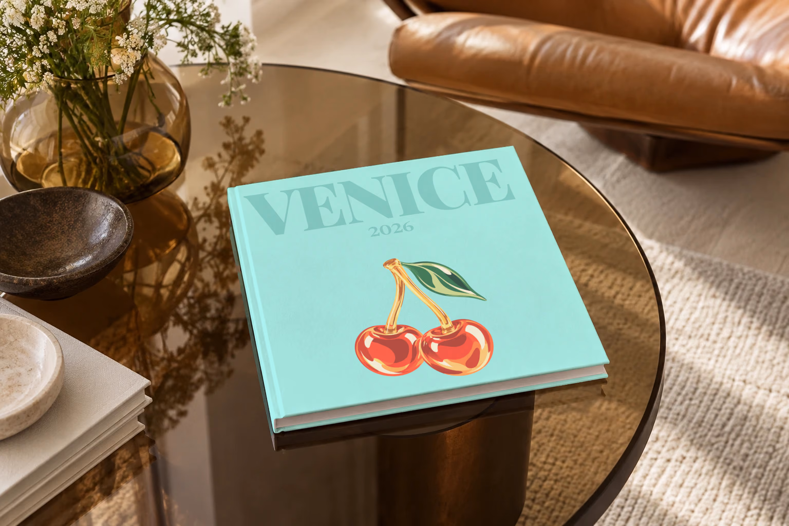 Venice square hardcover photo book with the illustrated cover of glass cherries on pale aqua, on a smoked glass coffee table beside a leather armchair in warm afternoon light