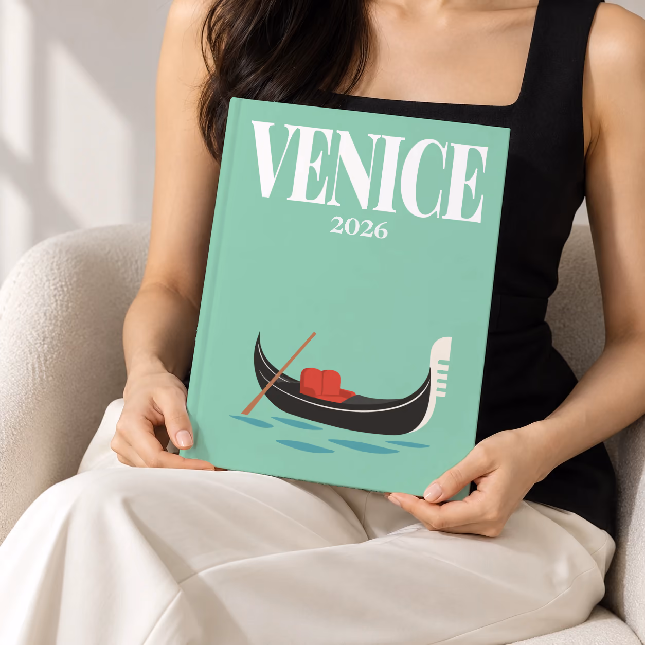 Venice hardcover photo book with the illustrated cover of a gondola on the lagoon on mint green, held upright in two hands by a woman in a black top, sitting in a cream boucle armchair