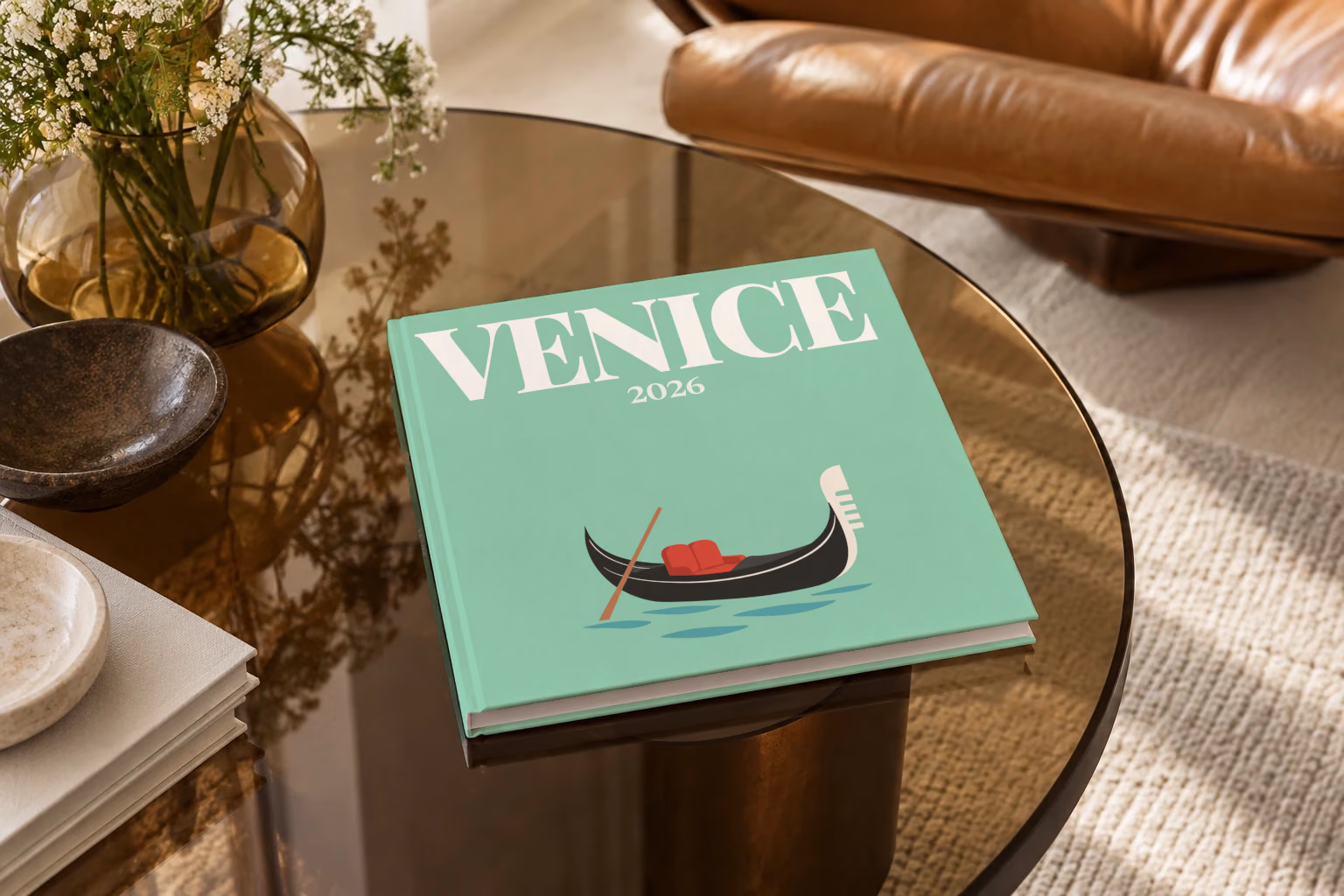 Venice square hardcover photo book with the illustrated cover of a gondola on the lagoon on mint green, on a smoked glass coffee table beside a leather armchair in warm afternoon light