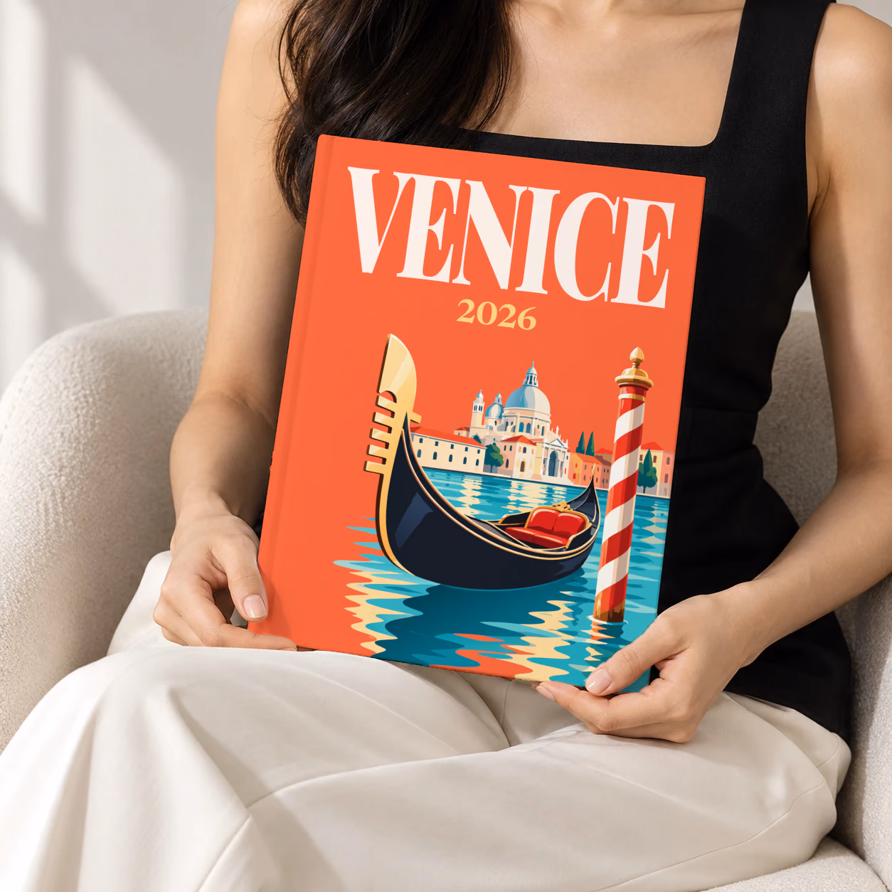 Venice hardcover photo book with the illustrated cover of a gondola, a striped mooring pole and Santa Maria della Salute on red-orange, held upright in two hands by a woman in a black top, sitting in a cream boucle armchair