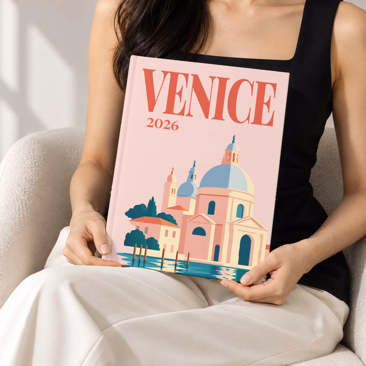 Venice hardcover photo book with the illustrated cover of Santa Maria della Salute on blush pink, held upright in two hands by a woman in a black top, sitting in a cream boucle armchair